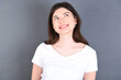 © Jihan - Portrait of mysterious young beautiful Caucasian woman wearing white T-shirt over studio grey wall looking up with enigmatic smile. Advertisement concept.