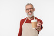 © InsideCreativeHouse - Smiling caucasian mature middle-aged man barista in apron holding to the camera selling coffee hot beverage tea drink in paper cup isolated in white background. Takeaway food