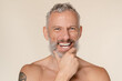 © InsideCreativeHouse - Smiling caucasian mature middle-aged man with bare shoulders naked shirtless with tattoos laughing looking at camera isolated in beige background.