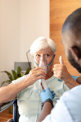 caucasian senior woman showing thumbs up while using oxygen mask sitting on a wheelchair at home