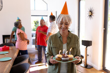 Asian Senior Woman Holding Cupcakes And Candles While Multiracial Friends Talking In Background
