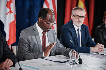 Wall Mural - African politician in eyeglasses sitting at table with microphone among his colleagues and reading report during press conference