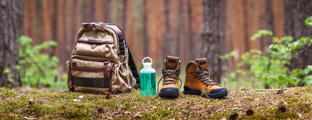 hiking and camping equipment in forest. backpack, water bottle and leather ankle boots. panoramic vi