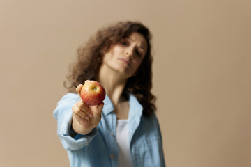 Wall Mural - Cute cheerful happy curly beautiful female in jeans casual shirt pulls apple at camera posing isolated on over beige pastel background. Healthy food. Natural eco-friendly products concept. Copy space