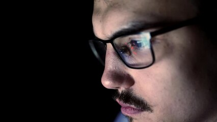 Closeup face of young man with glasses which reflects computer screen, front view. Man with glasses is sitting at computer in dark room