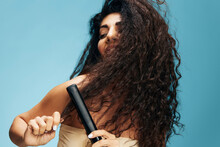 Joyful Happy Latin Curly Woman Using Hair Straightener, Looking Aside, Posing Isolated On Blue Wall Background. Hair Routine Concept, Haircare, Dry Damaged Hair Ironing, Hairdressing, Copy Place