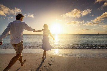 A romantic couple on holidays runs hand in hand on a tropical beach during sunset time