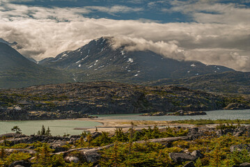 Wall Mural - Skagway, Alaska, USA - July 20, 2011: Klondike highway to Canada. Sandbank divides Summer Lake. Cloudscape over Canadian Rockies with snow patches and sparse green vegetation.