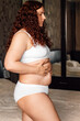 © Юля Бурмистрова - Curly plus size woman in white underwear standing near bed in bedroom and pressing on stomach with stretch marks closeup side view. Fat burning treatment of thick belly. Weight loss program, dieting.