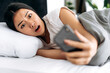Stunned chinese or japanese girl is lying in a bed in the morning, looks shocked at her smartphone, overslept the alarm clock, emotional facial expression, negative emotion, unexpected news or message