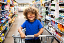 Funny Little Boy Laughing While Sitting In A Shopping Cart During A Family Trip To The Grocery Store. He Is Excited To Buy Something New	