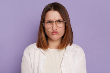 Portrait of Caucasian funny woman wearing white clothing and glasses, looking at camera with funny face and pout lips, posing isolated over purple background.