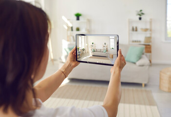 Person who plans on selling house or big spacious studio apartment takes photo on tablet device of clean bright Scandinavian Nordic home interior with stylish sofa, shelves, light walls, large windows