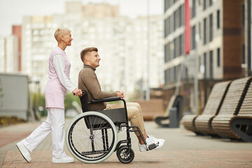 Content attractive qualified nurse in uniform strolling with disabled patient in city, she pushing wheelchair with man