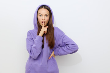 Wall Mural - a beautiful woman stands on a white background in a purple tracksuit, wearing a hood on her head, putting one hand on her belt, and leaning the other against her lips, looking forward in surprise