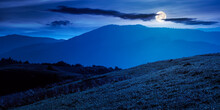 Mountainous Countryside Landscape At Night. Grassy Meadows And Trees On Hills Rolling In To The Distant Ridge In Full Moon Light