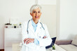 © Jelena Stanojkovic - Portrait of a mature female doctor standing in her office. Photo of a senior female doctor in a white coat with a stethoscope.