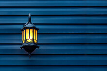 Iron Retro Lantern With Yellow Glass Shade Mounted On A Wooden Wall Of Blue Planks, Decorative Architecture Objects Of Evening Lighting With Warm Glow, Front View On Facade With Copy Space.