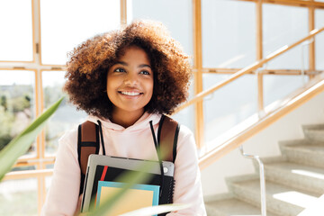 Wall Mural - Cheerful girl with backpack holding textbooks. Young female smiling and looking away in school.