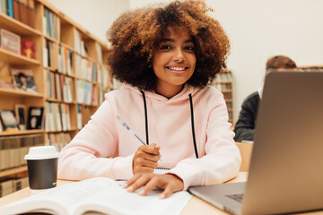 Wall Mural - Portrait of smiling young student studying in library. Young female at library desk looking at camera.