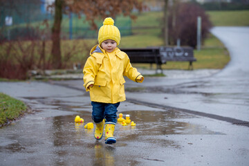 Poster - Beautiful funny blonde toddler boy with rubber ducks and colorful umbrella, jumping in puddles and playing in the rain