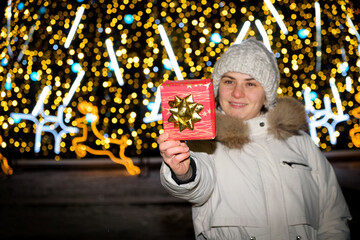 A woman in white winter clothes stands outside a Christmas tree, holds a red gift box and smiles.