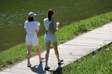 Two slim girls in beach shorts walking with cocktails in hands on wooden path on lake coast. Refreshing drinks at hot weather in summer city