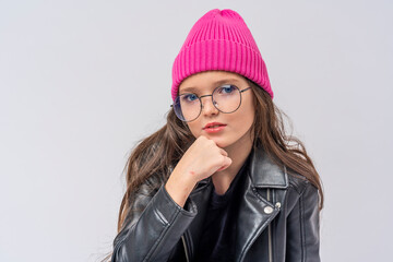 stylish teenage girl with dark hair, dressed in fashionable clothes, a leather jacket and a pink hat and glasses on white wall and looking at camera. transitional age. concept of youth and clothing.