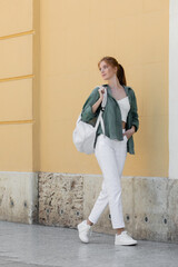 young redhead woman in green linen shirt holding backpack and walking near beige wall on street of valencia.