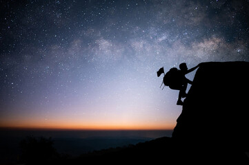Wall Mural - A young tourist with backpack is climbing a cliff alone with beautiful view of night sky, star and Milky Way. He had an effort to climb all the way to the top of the mountain and he had to succeed.