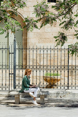 redhead woman sitting on concrete bench and using laptop near forged fence in valencia.