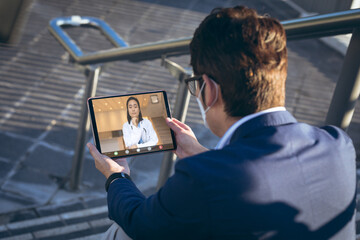 Sticker - Caucasian businessman wearing mask while sitting on steps during video call with asian female doctor