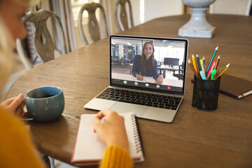 Poster - Happy caucasian businesswoman writing during video call with caucasian female colleague having drink