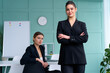 © Andrii - Two young women leaders dressed black suit in office one woman standing arms crossed other woman sitting on the table looking at camera. Business meeting