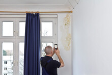 Man Taking A Photo Of A Big Patch Of Mold Or Mildew Growing Behind The Drapes Of A White External Wall In An Old House.