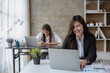 © aksonsat - Young business women working on laptop and writing on note book with happy and smile face with co-worker is talking via mobile phone behind her in office spec.