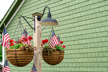 Patriotic Hanging Baskets In Front Of Green Building With United States Flags, Geraniums And Sweet Potato Plant In Black Metal Wire Basket With Coco Coir Liners