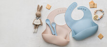 Silicone Baby Bibs With Plastic Cutlery And Toys On Light Background With Space For Text