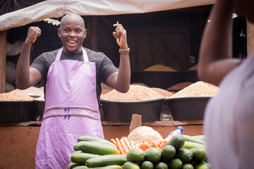 An African Nigerian male trader, seller, business man or shop owner, having an apron on his body, happily jubilating as he holds a smart phone in a market