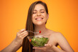 Happy woman closed eyes with bare shoulders and long hair holding bowl with green salad and tomatoes isolated on orange yellow background.