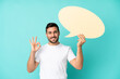 canvas print picture - Young handsome caucasian man isolated on blue background holding an empty speech bubble and doing OK sign