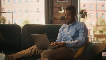 Leinwandbilder - Young Handsome Black Man Sitting on Couch in Loft Living Room, Working from Home on Laptop Computer. Creative Male Pondering while Checking Social Media and Browsing Internet