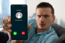 Upset Man Holding A Smartphone With Unknown Caller Displayed On Screen. Unknown Caller Telephony Concept. Incoming Call At Mobile Phone. Prank Caller, Scammer Or Stranger, Fraud, Cybercrime.