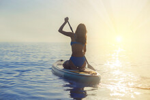 Asian Beautiful Girl In A Blue Bikini Is Engaged In Water Sports. She Sits With Her Back On A Surfboard With A Paddle In The Ocean Against The Background Of A Sunset. Woman On Paddleboarding , Sup.