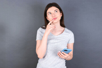 young beautiful Caucasian woman wearing white T-shirt over grey wall thinks deeply about something, uses modern mobile phone, tries to made up good message, keeps index finger near lips.