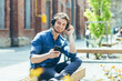 © Liubomir - Young man, student guy sitting on campus on a bench in the lotus position, listening to an audiobook, audio lecture in headphones, holding a phone in his hands. He smiles.