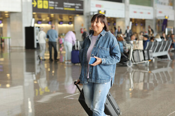 smiling middle aged woman with suitcase and passport standing in arrival or departure hall of an airport. Holiday vacation.