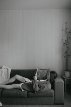 Black And White Photo Of A Girl On The Couch, Against A Light Wall, Reading A Book