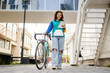 © Miljan Živković - One woman young adult caucasian female walking by the university building holding bicycle and digital tablet in day real people copy space happy joyful full length front view