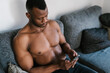 © Ezequiel Giménez/Stocksy - Shirtless black man messaging on smartphone on couch